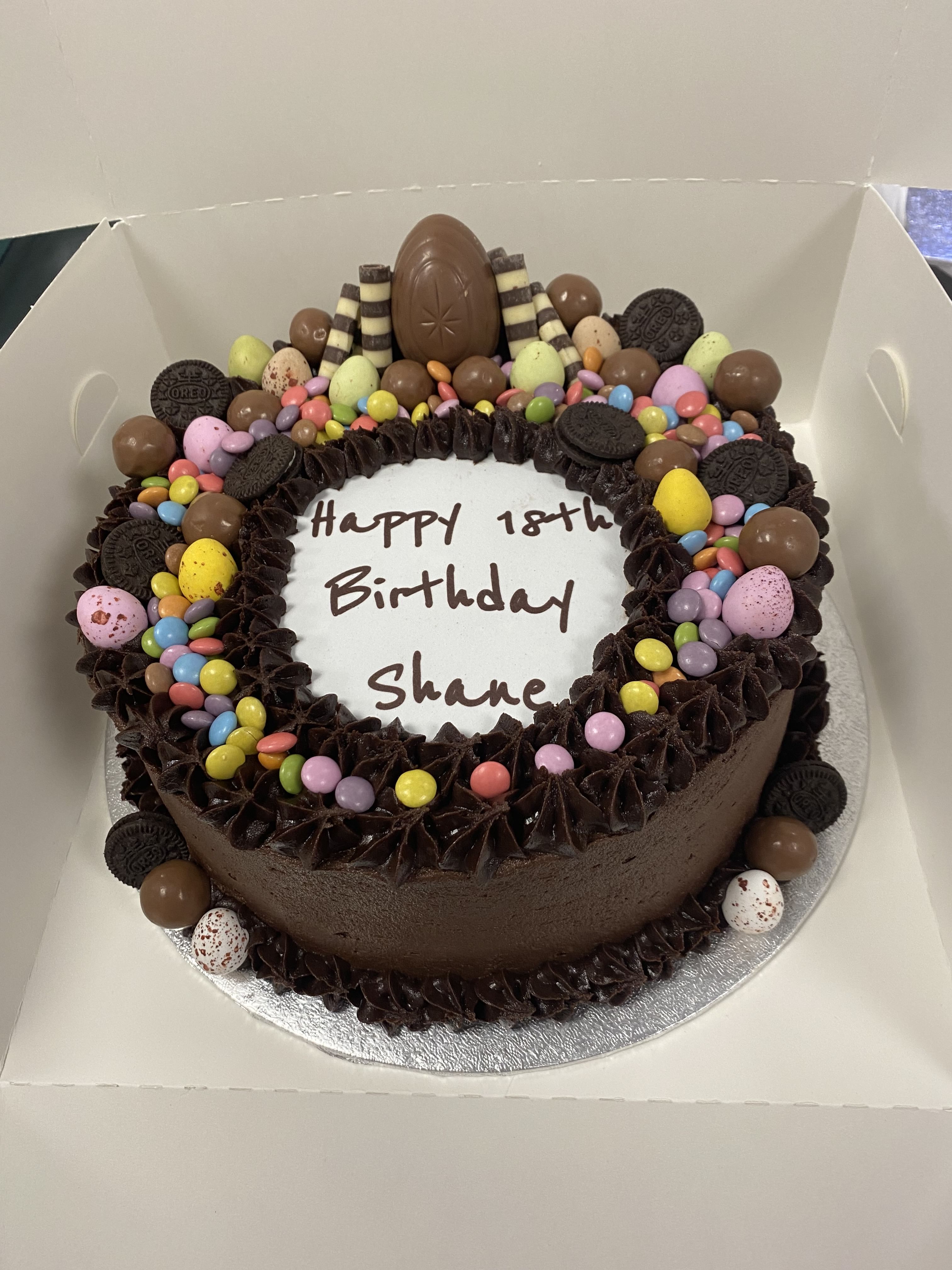 A round chocolate birthday cake, adorned with various colorful candies, chocolate eggs, and cookies, boasts a rich layer of chocolate frosting. An oval chocolate centerpiece sits on top. The cake reads 'Happy 18th Birthday Shane' in the center and is presented in an open white box—a true crowd-pleasing dessert.